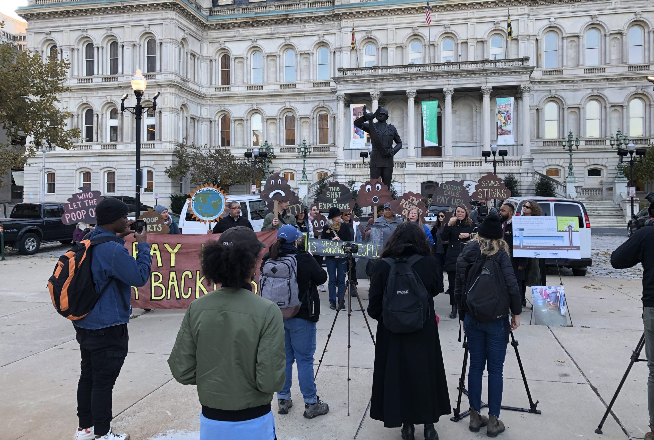 A group protests outside city hall in Baltimore, as news cameras look on.
