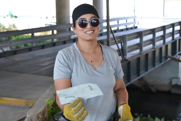 Pictured: A woman wearing sunglasses, a grey Under Armour shirt, and yellow gloves holding an unsighed check for $75 found at a cleanup site at Cheers to Clean Water 2019.