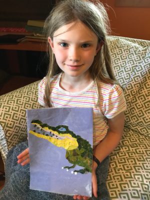 A young girl holding up a completed nature collage of an alligator.