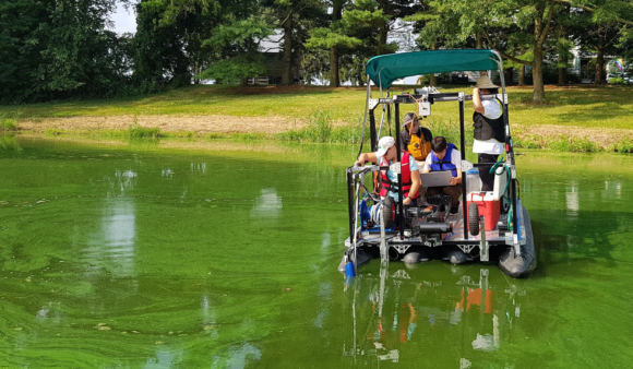 Matt Stocker and three USDA scientists on a silver pontoon boat in a shallow green colored water body.