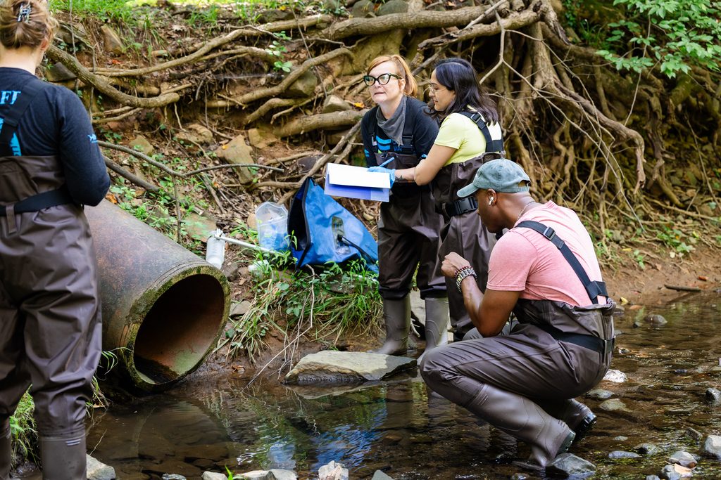 Outfall Screening blitz at a large stormpipe. Several water experts and volunteers take notes, analyze trash, and collect water samples. They all wear t shirts with waders and high boots.
